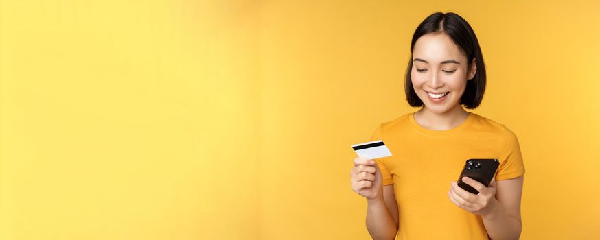 Online shopping. Smiling asian girl using credit card and mobile phone app, paying contactless, order on smartphone application, standing over yellow background.