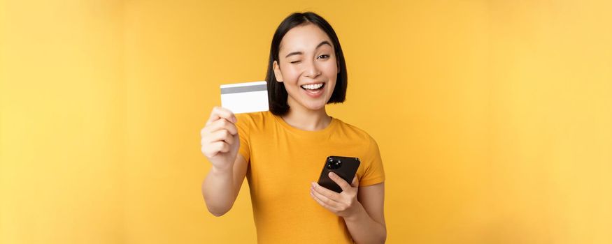 Joyful asian girl smiling, showing credit card and smartphone, recommending mobile phone banking, standing against yellow background.