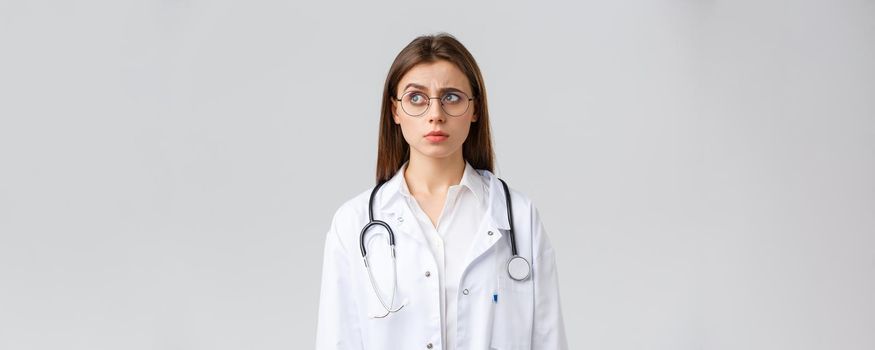 Healthcare workers, medicine, insurance and covid-19 pandemic concept. Doctor see something strange. Confused and suspicious female doctor in white scrubs, glasses, frowning and staring left.