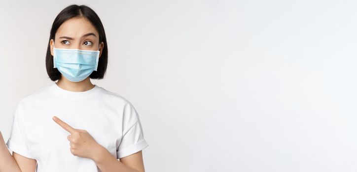 Image of asian woman in medical mask from covid, looking confused left, pointing at logo, showing advertisement, standing over white background.