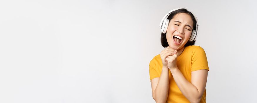 Happy asian girl dancing, listening music on headphones and smiling, standing in yellow tshirt against white background. Copy space