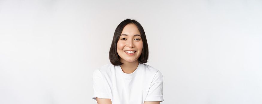 Beautiful korean girl smiling, white teeth, looking lovely at camera, standing in white tshirt over studio background.