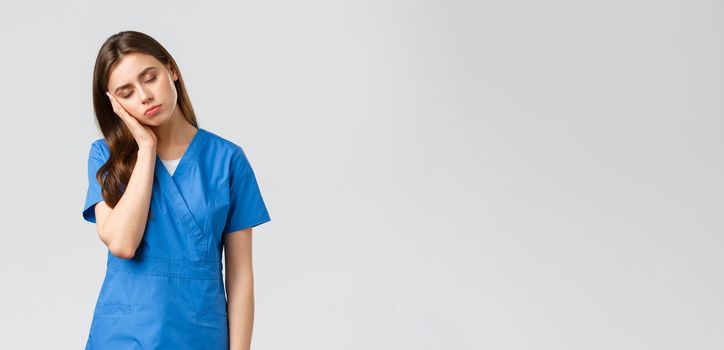 Healthcare workers, prevent virus and medicine concept. Tired sleepy female doctor, young nurse in blue scrubs, lean on palm and close eyes, exhausted from night shift at clinic fighting coronavirus.