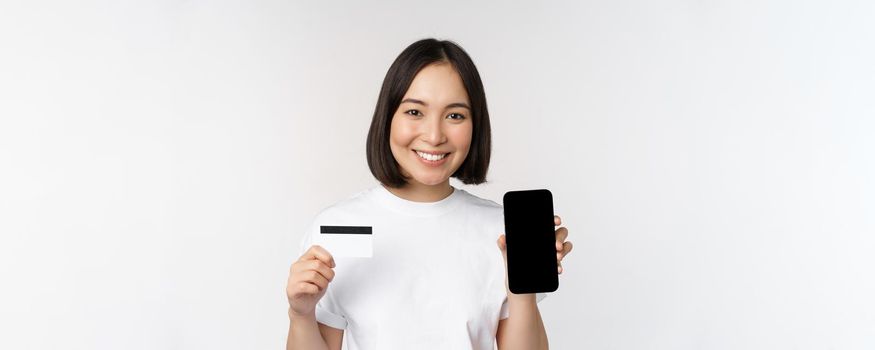 Portrait of smiling young asian woman showing mobile phone screen and credit card, standing over white background.