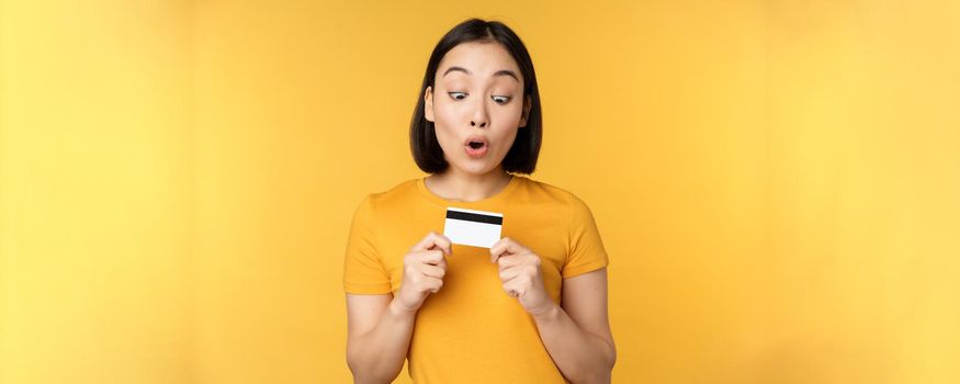 Beautiful asian woman showing credit card and smiling, recommending bank service, standing over yellow background. Copy space