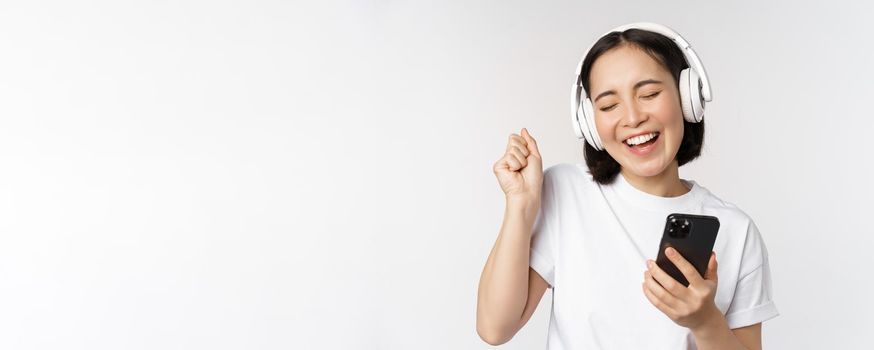 Beautiful modern asian girl, listening music in headphones, holding mobile phone, using smartphone app, standing over white background.