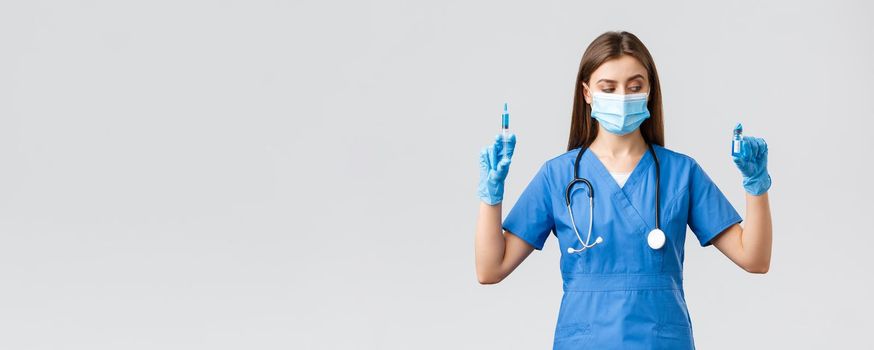 Covid-19, preventing virus, health, healthcare workers and quarantine concept. Female nurse or doctor in blue scrubs, medical mask and gloves, look at ampoule with coronavirus vaccine, hold syringe.