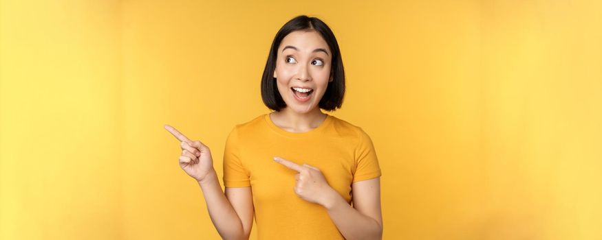 Portrait of happy asian girl pointing fingers and looking left, smiling amazed, checking out promo banner, showing advertisement against yellow background.