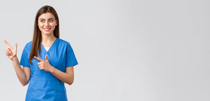 Healthcare workers, prevent virus, insurance and medicine concept. Smiling pretty nurse or doctor in blue scrubs, pointing fingers left and looking at baner with pleased grin, grey background.