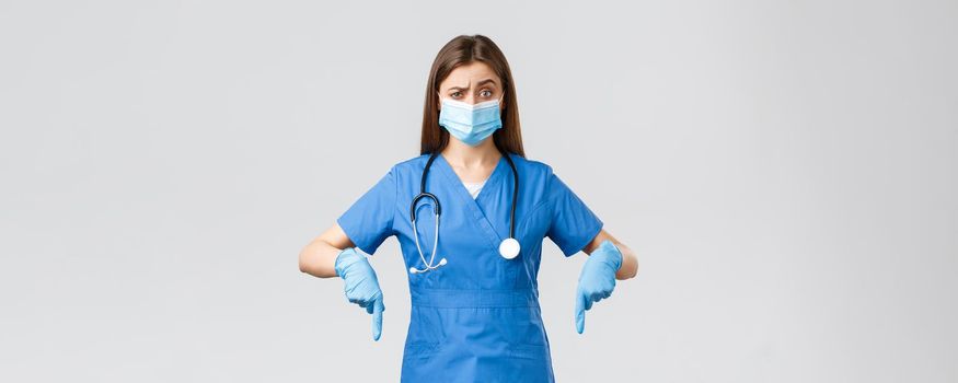 Covid-19, preventing virus, health, healthcare workers and quarantine concept. Skeptical and unsure female nurse or doctor in blue scrubs, personal protective equipment, pointing fingers down unsure.