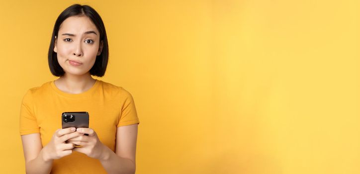 Skeptical asian woman holding smartphone, looking with doubt at camera, standing over yellow background.