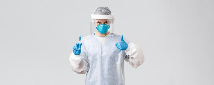 Covid-19, medical research, healthcare workers and quarantine concept. Confident professional doctor in personal protective equipment PPE costume, show thumb-up, hold syringe with corona vaccine.