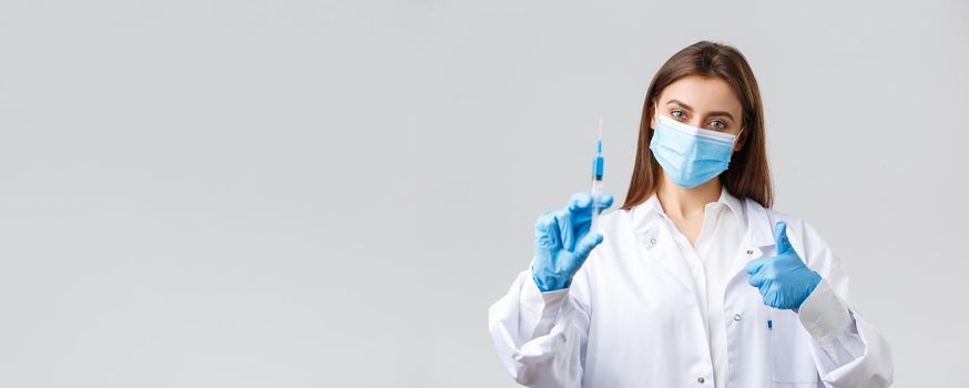 Covid-19, preventing virus, healthcare workers and quarantine concept. Determined young doctor in medical mask and rubber gloves, scrubs show thumbs-up and syringe with coronavirus vaccine.