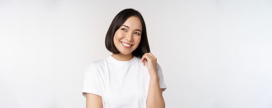 Portrait of cute coquettish woman laughing and smiling, looking aside thoughtful, thinking or imaging smth, standing in white t-shirt over studio background.