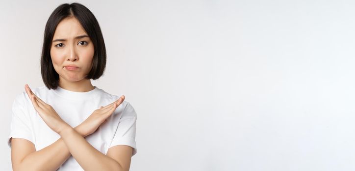 Portrait of asian korean woman showing stop, prohibition gesture, showing arm cross sign, standing in tshirt over white background. Copy space