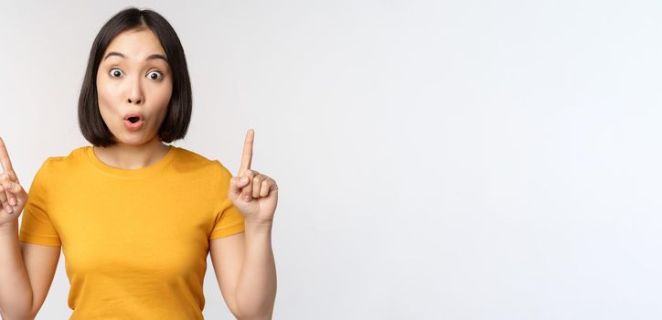Portrait of impressed asian girl, pointing fingers up, say wow, looking amazed at camera, standing over white background. Copy space