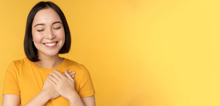 Beautiful asian woman, smiling with tenderness and care, holding hands on heart, standing in tshirt over yellow background.