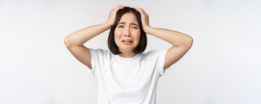 Desperate young korean woman holding hands on head, panicking, crying and standing distressed against white background.