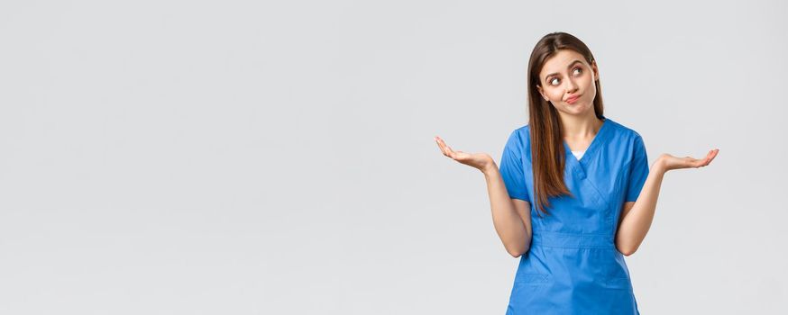 Healthcare workers, prevent virus, insurance and medicine concept. Indecisive and clueless attractive doctor, female nurse in blue scrubs, shrugging with hands sideways and smirk, look away.