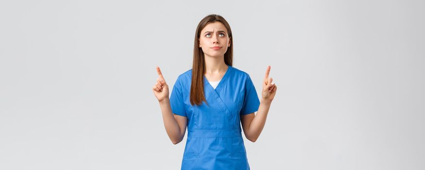 Healthcare workers, prevent virus, covid-19 test screening, medicine concept. Dissatisfied and upset young moody nurse, doctor in blue scrubs, smirk and frowning, pointing finger up.