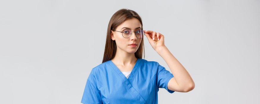 Healthcare workers, medicine, insurance and covid-19 pandemic concept. Professional smart female nurse, doctor or intern in blue scrubs and glasses looking at camera determined, grey background.