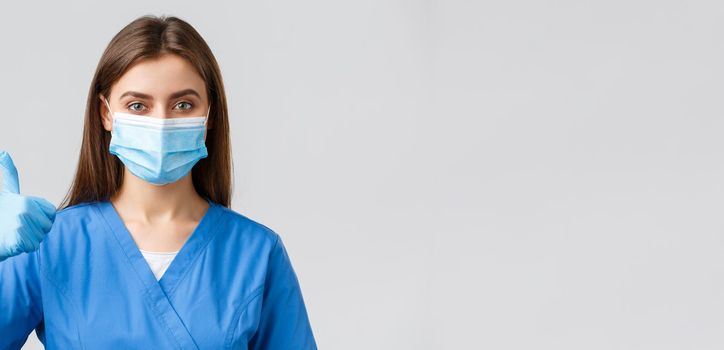 Covid-19, preventing virus, health, healthcare workers and quarantine concept. Confident supportive female nurse or doctor in blue scrubs, medical mask, thumbs-up, approve or recommend.