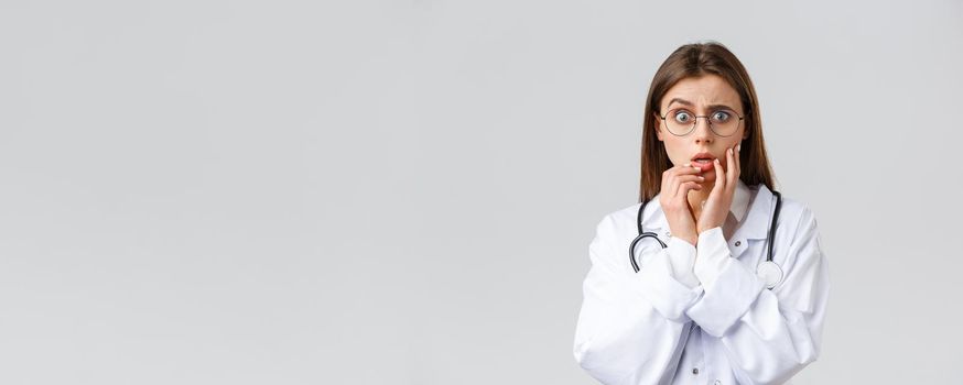 Healthcare workers, medicine, insurance and covid-19 pandemic concept. Shocked scared and concerned female doctor in white scrubs and glasses listening to scary news, gasping, hold hands near lips.