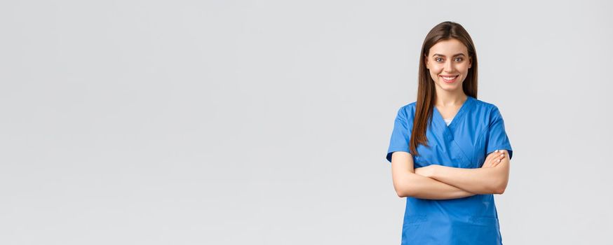 Healthcare workers, prevent virus, insurance and medicine concept. Confident female doctor, nurse in blue scrubs, smiling and cross arms chest, ready to save patient lives, grey background.