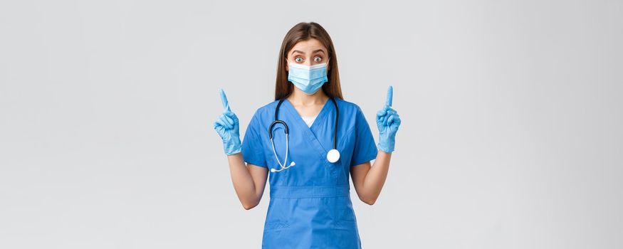 Covid-19, preventing virus, health, healthcare workers concept. Surprised or curious female nurse in blue scrubs, personal protective equipment, pointing fingers up, found out interesting news.