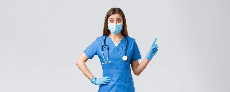 Covid-19, preventing virus, health, healthcare workers concept. Intrigued and excited female doctor, nurse in blue scrubs and medical mask inform patients, pointing fingers upper right corner.