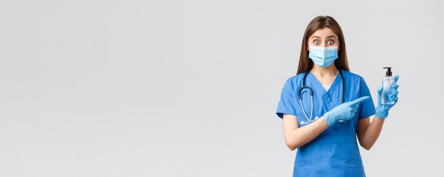 Covid-19, preventing virus, health, healthcare workers and quarantine concept. Enthusiastic female nurse or doctor in blue scrubs, medical mask and gloves, pointing at hand sanitizer, look surprised.
