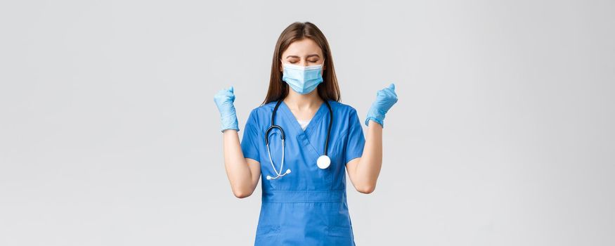 Covid-19, preventing virus, health, healthcare workers and quarantine concept. Encouraged happy female nurse or doctor in blue scrubs, celebrating, wear medical mask, chanting yes in success.