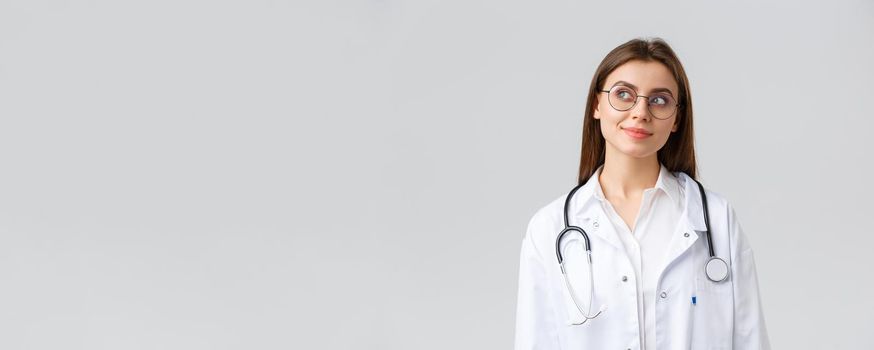 Healthcare workers, medicine, insurance and covid-19 pandemic concept. Dreamy young talented doctor in white scrubs and glasses, looking upper left corner thoughtful and smiling, imaging something.