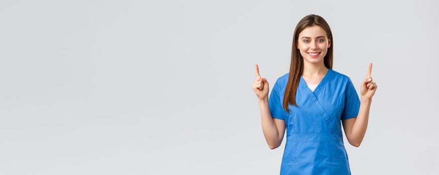 Healthcare workers, prevent virus, insurance and medicine concept. Attractive nurse or doctor in blue scrubs, pointing fingers up and smiling, advertise banner, medical pills, grey background.