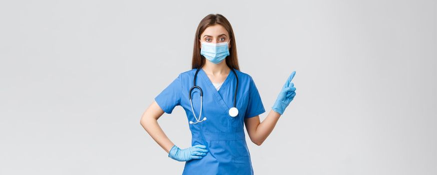 Covid-19, preventing virus, health, healthcare workers and quarantine concept. Confident female nurse in blue scrubs and medical mask provide information, pointing fingers right.