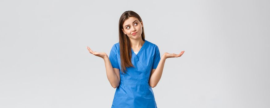 Healthcare workers, prevent virus, insurance and medicine concept. Indecisive and clueless attractive doctor, female nurse in blue scrubs, shrugging with hands sideways and smirk, look away.