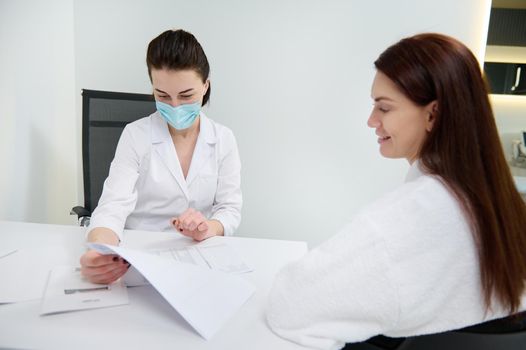 Confident doctor, gemeral practictioner, aesthetician consulting her patient, analyzing her medical records in a white office. Concept of a trusting relationship with doctor. Health care and medicine