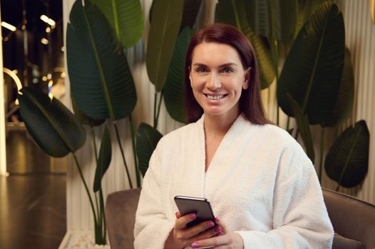 Attractive European woman in white bathrobe, sits in armchair in luxury spa lounge zone, smiles toothy smile looking at camera and checks her mobile phone while relaxing in cosmetology wellness centre