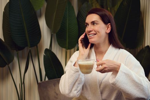 Delightful woman holding a cup with cappuccino or coffee drink and talking on telephone while relaxing in the spa lounge after receiving rejuvenating anti-aging treatment. Cosmetology, spa concept