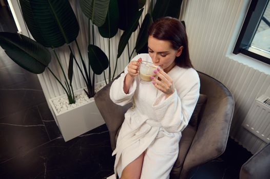 Top view of an attractive middle aged European woman with perfect clean fresh skin in white terry bathrobe drinking cappuccino in the lounge cafe of a wellness spa clinic and smiling looking at camera