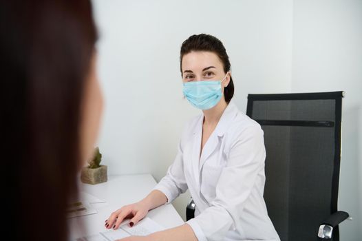 Confident portrait of a brunette Caucasian woman, female doctor in medical mask looking at camera while consulting a patient in the modern minimalist medical office in a clinic with white interior