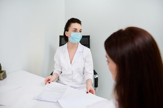 Rear view. Woman at medical appointment, sitting in front of a general practitioner, receiving consultation in a clinic with minimalist white interior. Cosmetology, health care and medicine concept