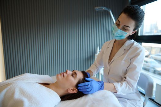 Relaxed elegant Caucasian middle-aged woman enjoying a professional massage on her head done by a massage therapist while lying on a couch in a wellness spa center.