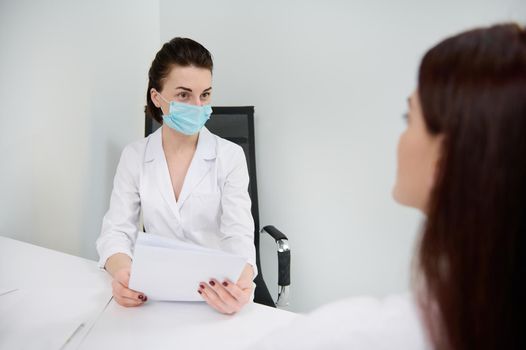 Confident female doctor general practitioner esthetician cosmetologist in protective medical mask consults a patient during regular medical check-up in a modern clinic with minimalist white interior