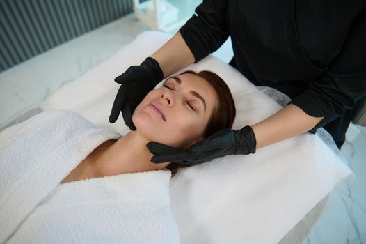 Overhead view of charming elegant middle aged European woman enjoying professional massage on her face, lying on a couch while a masseuse performing lymphatic drainage and anti-aging facial treatment