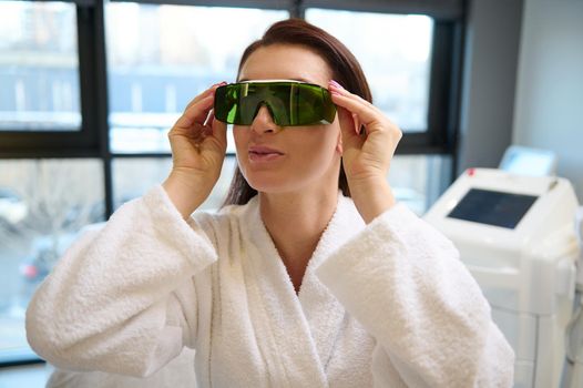 Confident European middle aged woman in white bathrobe puts on protective UV eyeglasses, ready for beauty treatment with laser apparatus in a wellness spa centre with contemporary medical equipments.