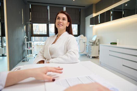 Charming Caucasian woman in a white bathrobe talking to cosmetologist doctor, consulting about anti-aging treatment procedures, in a cosmetology clinic with modern equipment for professional body care