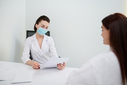 Confident female doctor in medical mask consults her patient, analyzes her medical records in a white office. Concept of a trusting relationship with doctor. Health care and medicine. Cosmetology