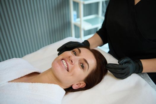 Happy delightful charming middle aged dark-haired Caucasian woman smiling with cheerful toothy smile while lying on a massage couch, enjoying beauty and wellness therapy in modern spa salon