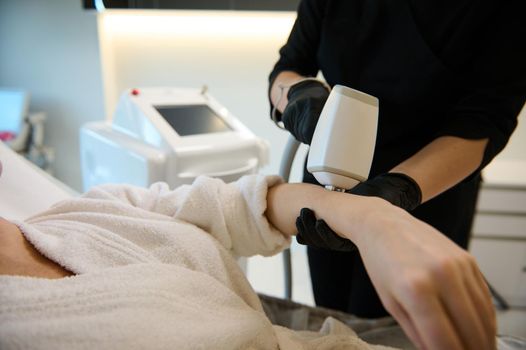 Close-up of an unrecognizable cosmetologist aesthetician performing a hair removal procedure on the hand of a woman with modern laser equipment in wellness spa clinic